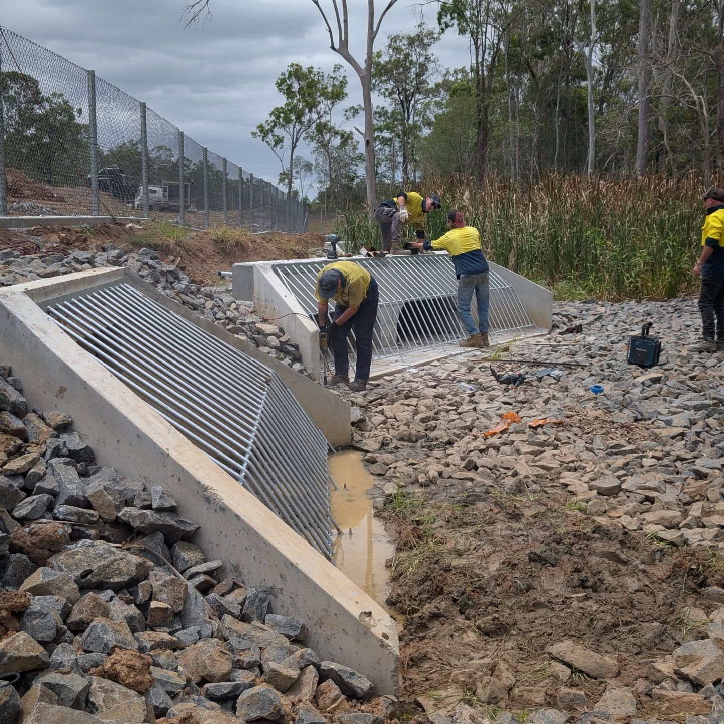 Workers installing concrete drainage grates in a rocky roadside ditch near a chain-link fence