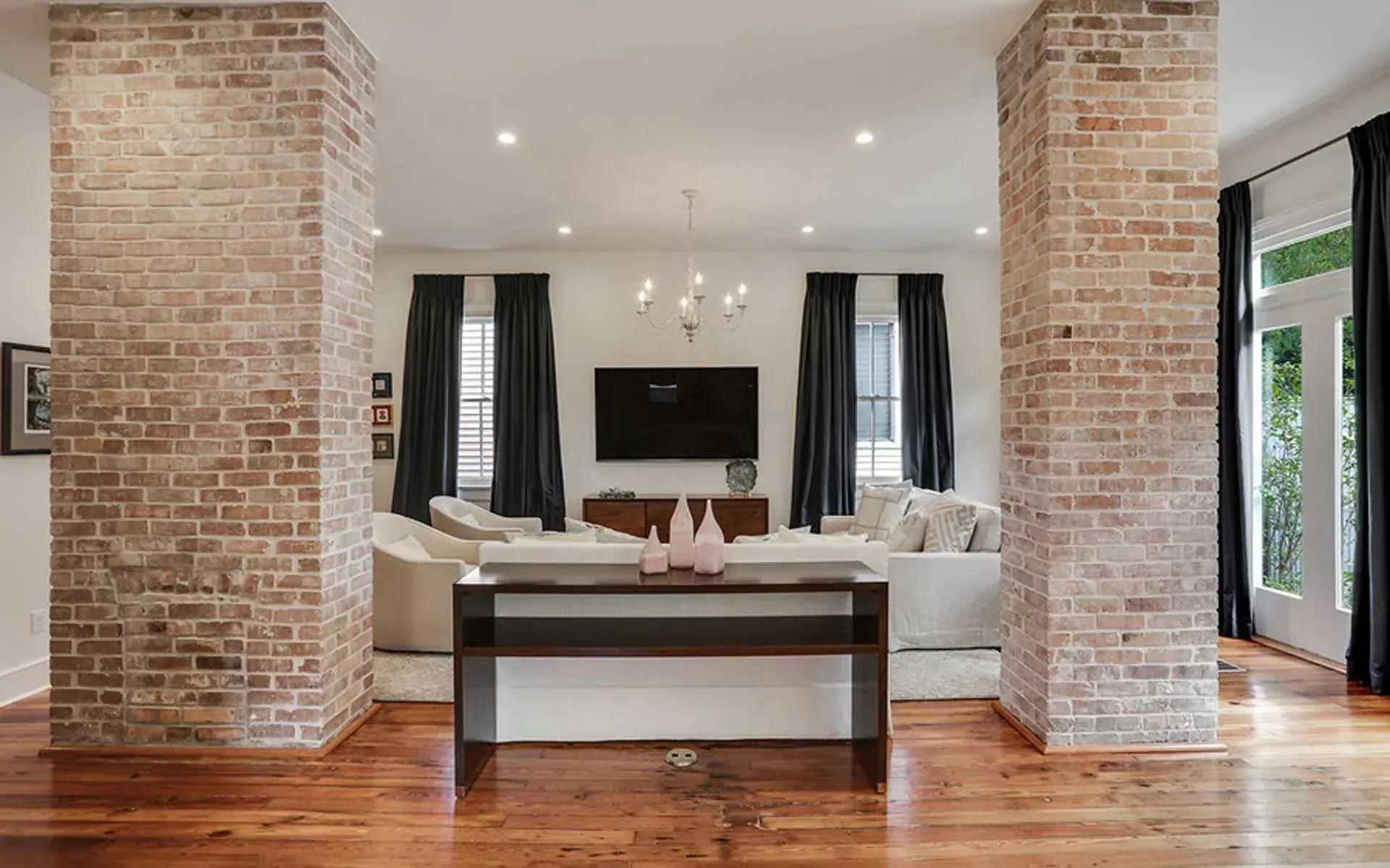 Living room with exposed brick pillars, hardwood floors, white furniture, and black curtains.