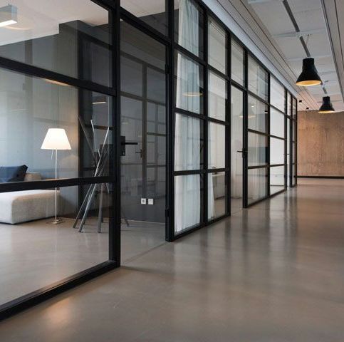 Modern office interior with glass-walled rooms, dark frames, gray floor, white couch, and pendant lights.