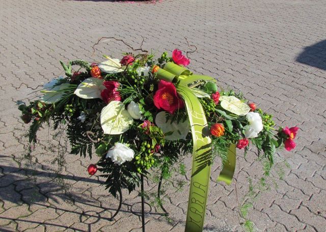 Composizione floreale funebre con anthurium bianchi e fiori rossi, impreziosita da un nastro verde, su un supporto metallico.