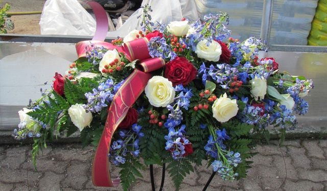 Composizione floreale per bara con rose rosse e bianche, delphinium blu, verde e un vistoso nastro rosso su un supporto.