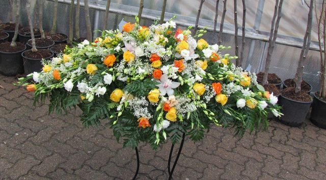 Una grande composizione floreale per bara, con rose gialle e arancioni, gigli bianchi e verde, su un supporto in un vivaio.