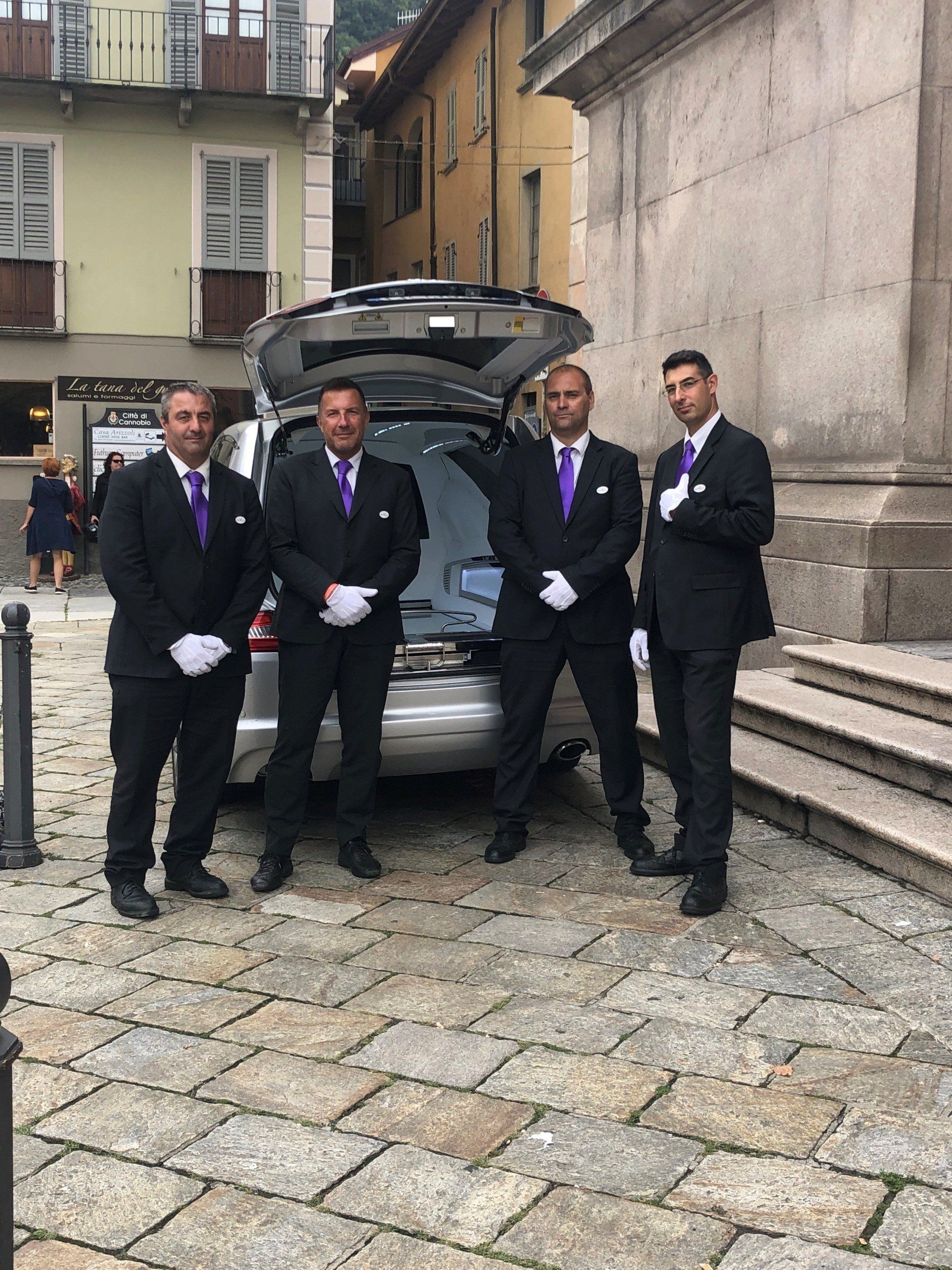 Quattro uomini in abiti neri coordinati, guanti bianchi e cravatte viola sono in piedi, in posizione formale