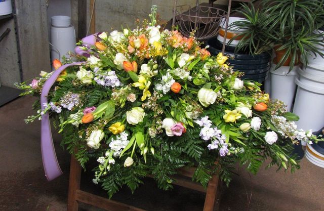 Una grande composizione floreale funebre di fiori bianchi, arancioni e viola con verde, poggiata su un supporto di legno.