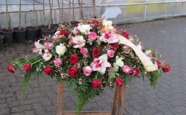 Composizione floreale funebre con rose rosse e rosa, gigli e verde, disposta su un supporto di legno all'aperto.