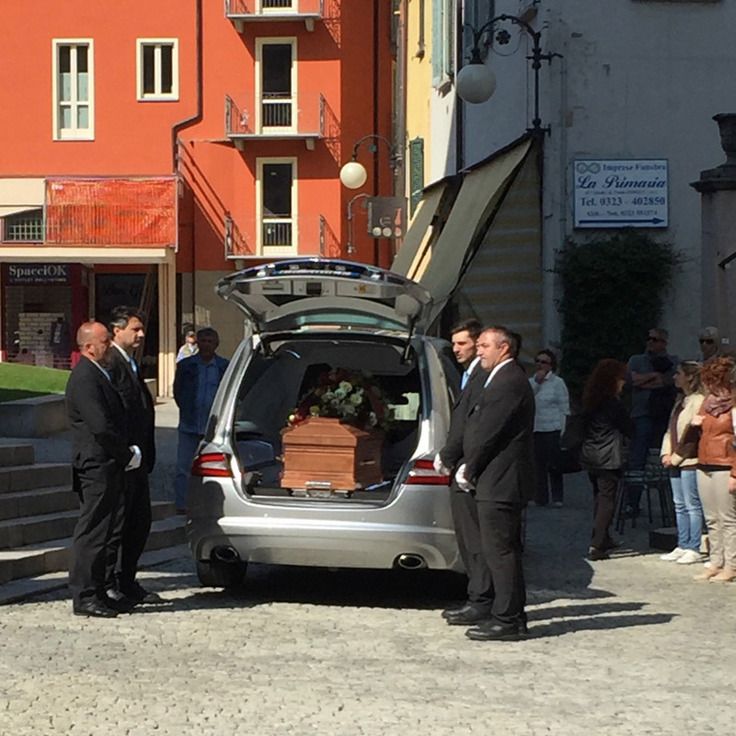 Quattro persone in abito elegante sono in piedi vicino a un carro funebre con il retro aperto, contenente una bara di legno