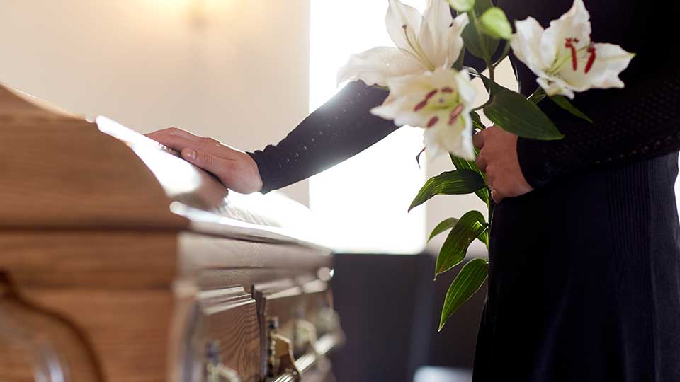 Durante un funerale, una persona vestita di nero tiene in mano dei gigli bianchi e appoggia la mano su una bara di legno.