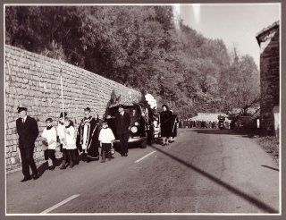Una fotografia in bianco e nero di un corteo funebre che percorre una strada accanto a un muro di pietra