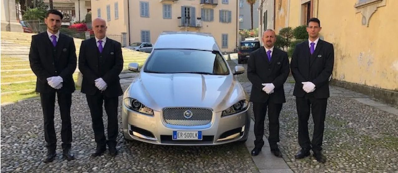 Quattro individui in abiti formali neri e guanti bianchi sono in piedi ai lati di un carro funebre argentato