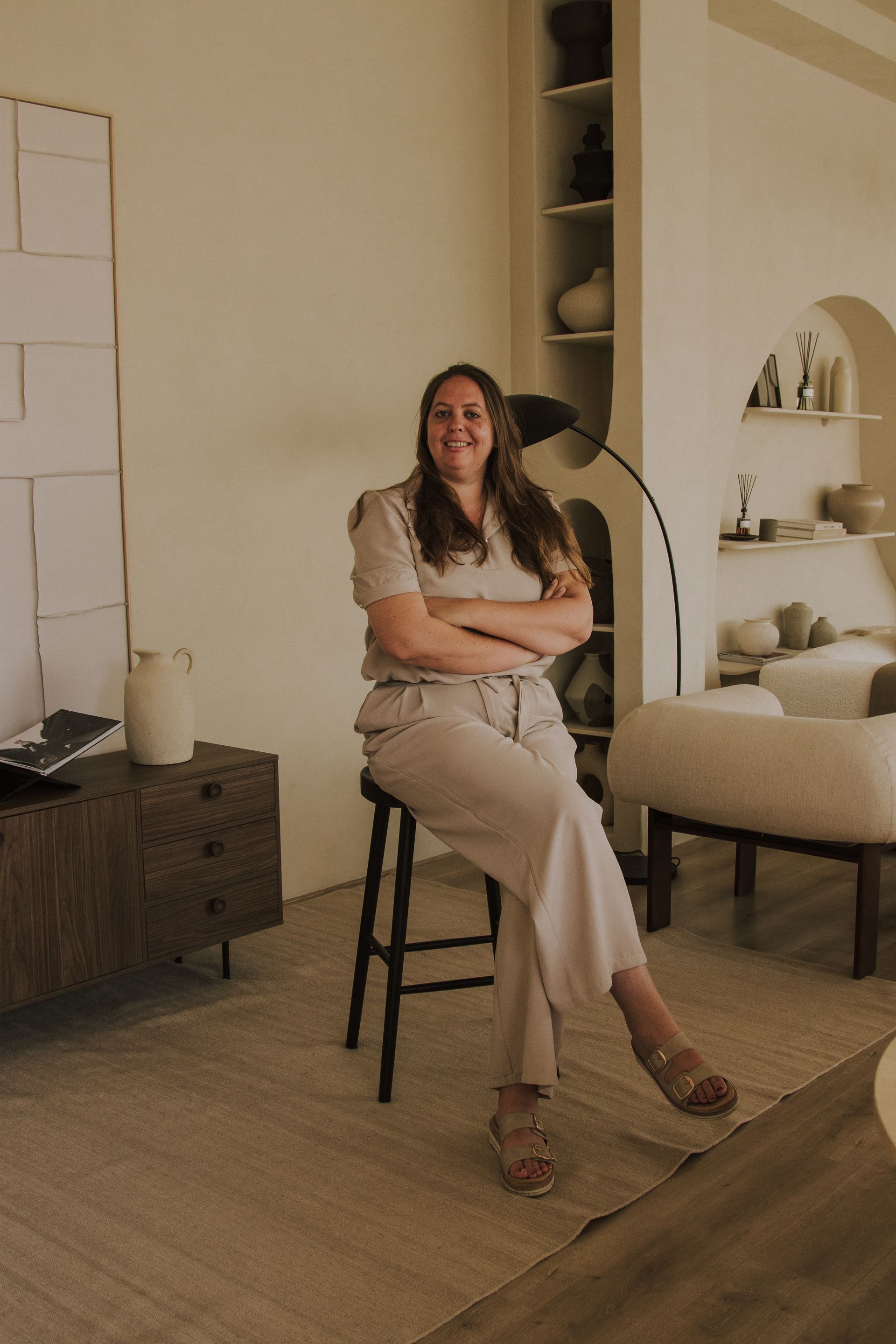 Vrouw zit op een krukje in een gezellige, beige woonkamer en glimlacht met haar armen over elkaar.