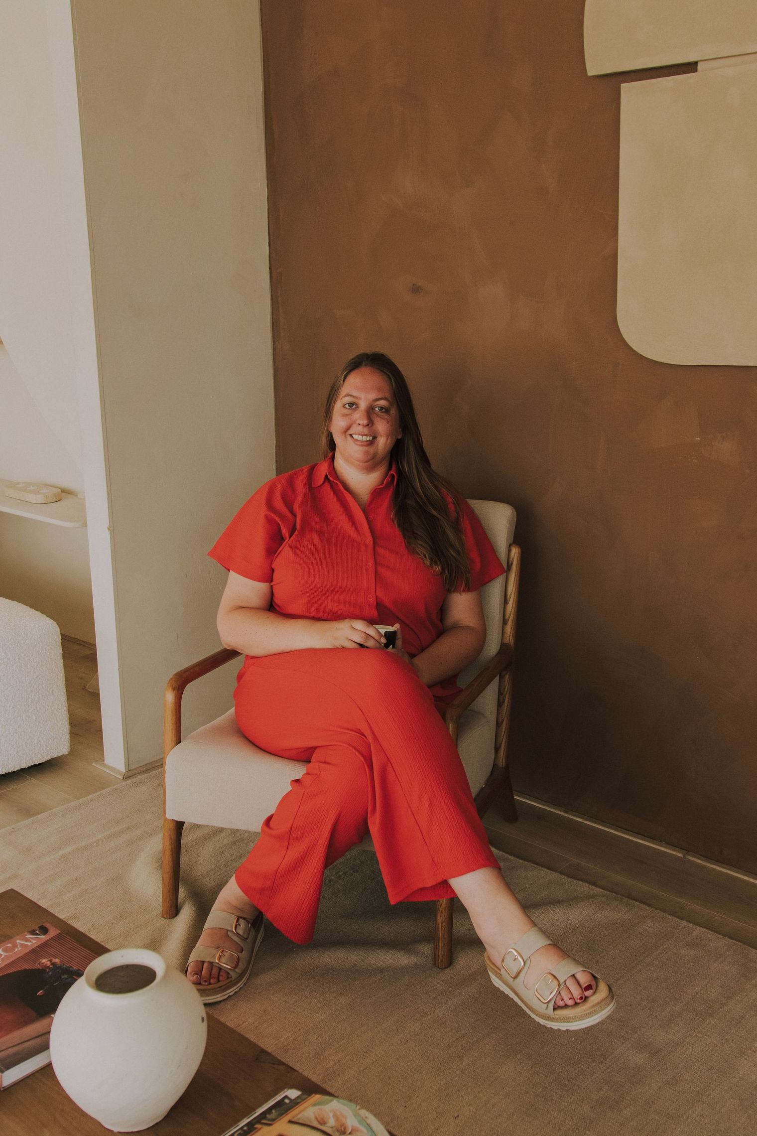 Een persoon in een rode outfit zit met gekruiste benen op een stoel en glimlacht in een moderne kamer.