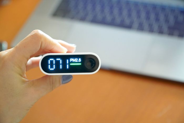 Hand holding a white air quality monitor showing a PM2.5 reading of 071, with a laptop in the background.