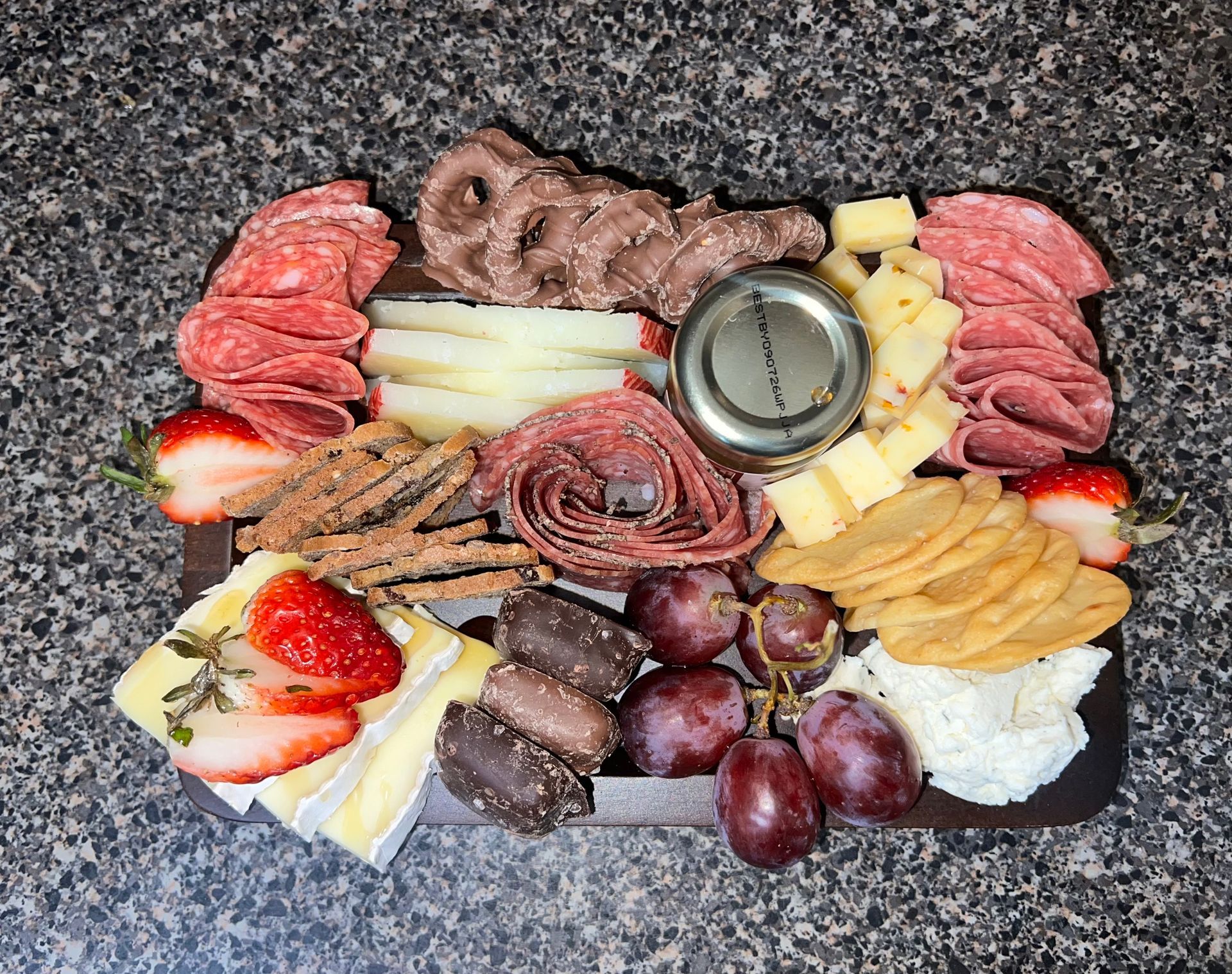 Assorted charcuterie board with meats, cheeses, strawberries, grapes, and a central dip on a dark countertop