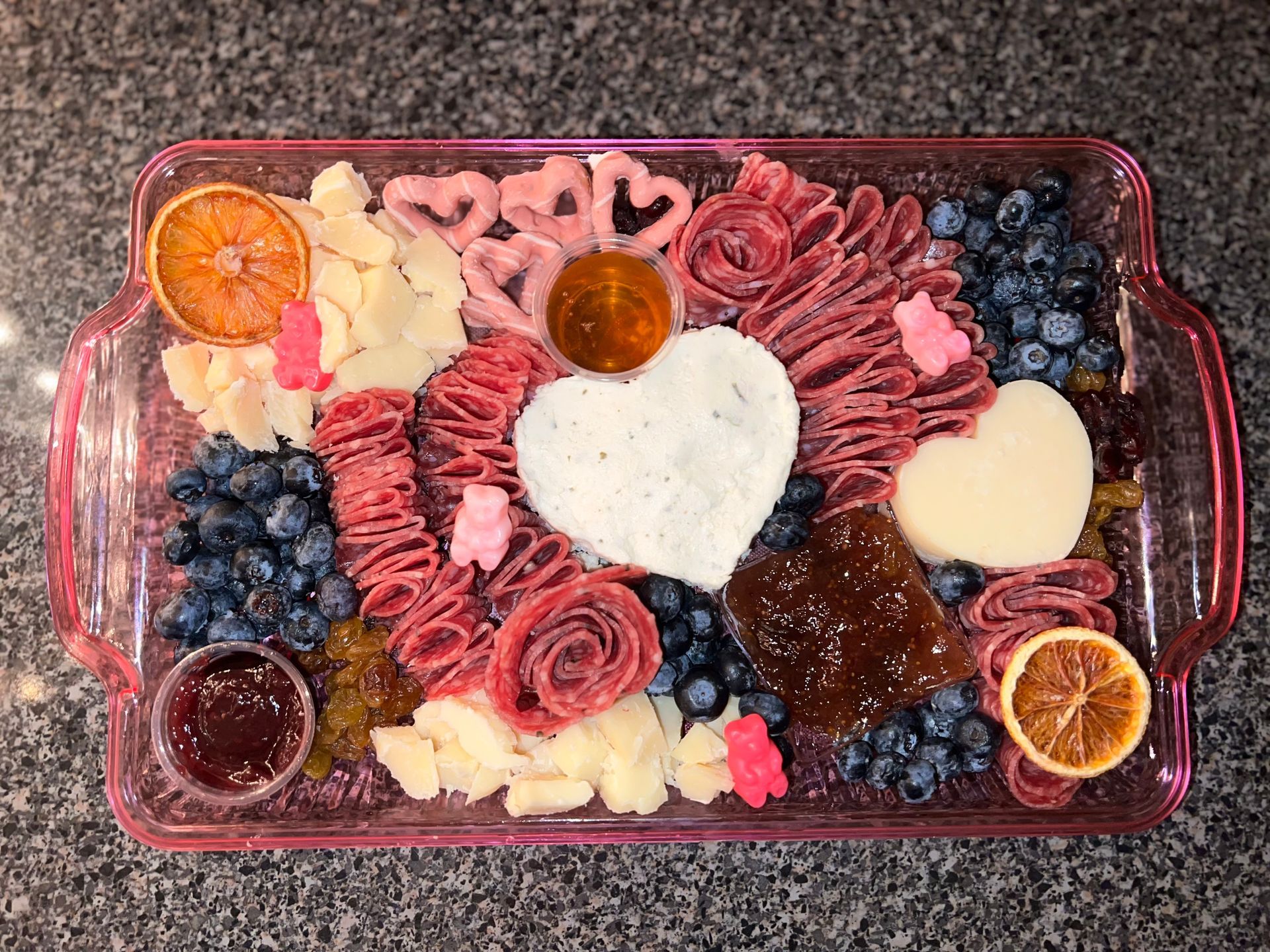 Heart-shaped charcuterie board with cheeses, salami roses, grapes, berries, and crackers on a dark tray