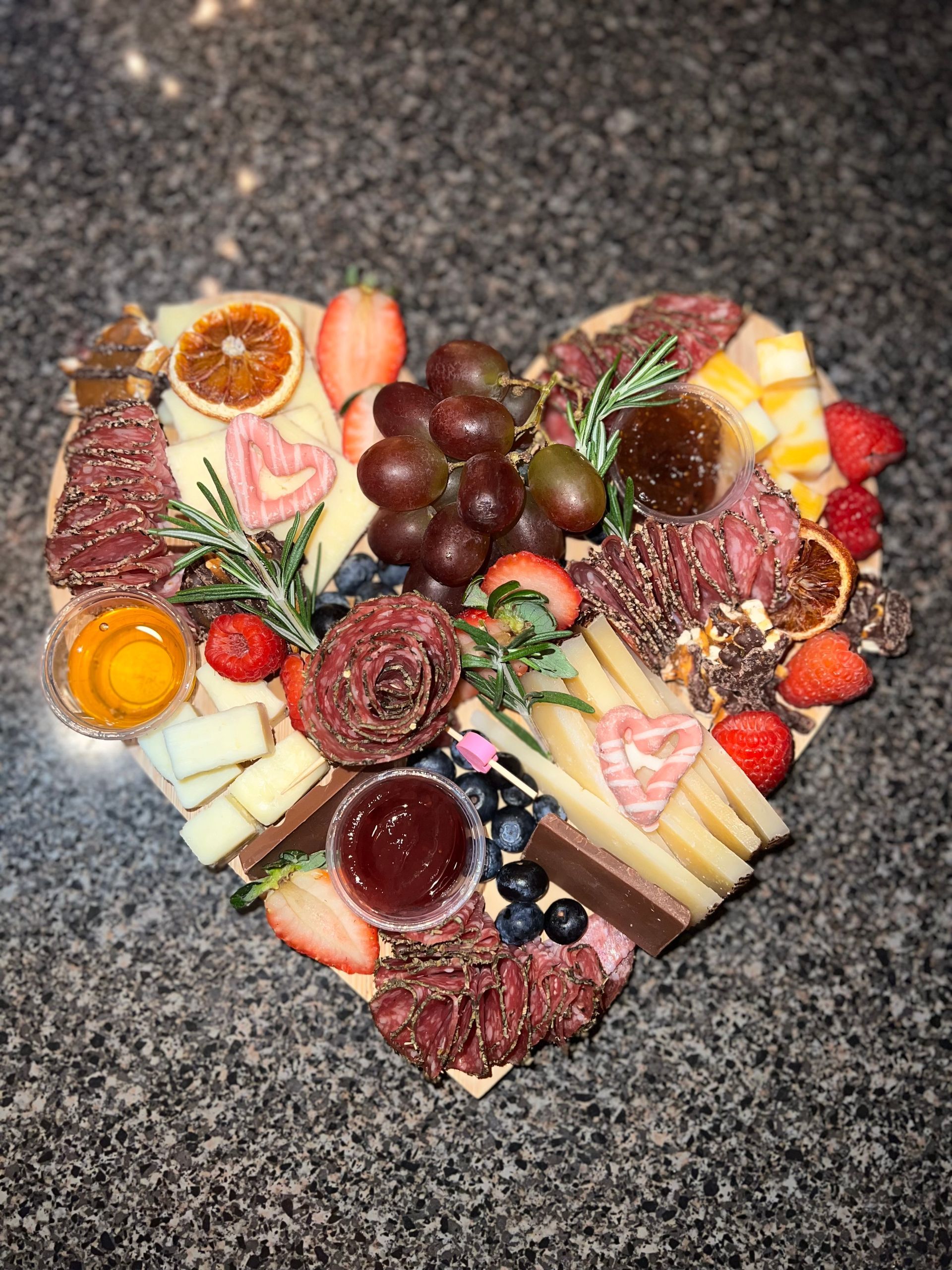 Heart-shaped charcuterie board with fruit, cheese, crackers, meats, and grapes on a dark countertop