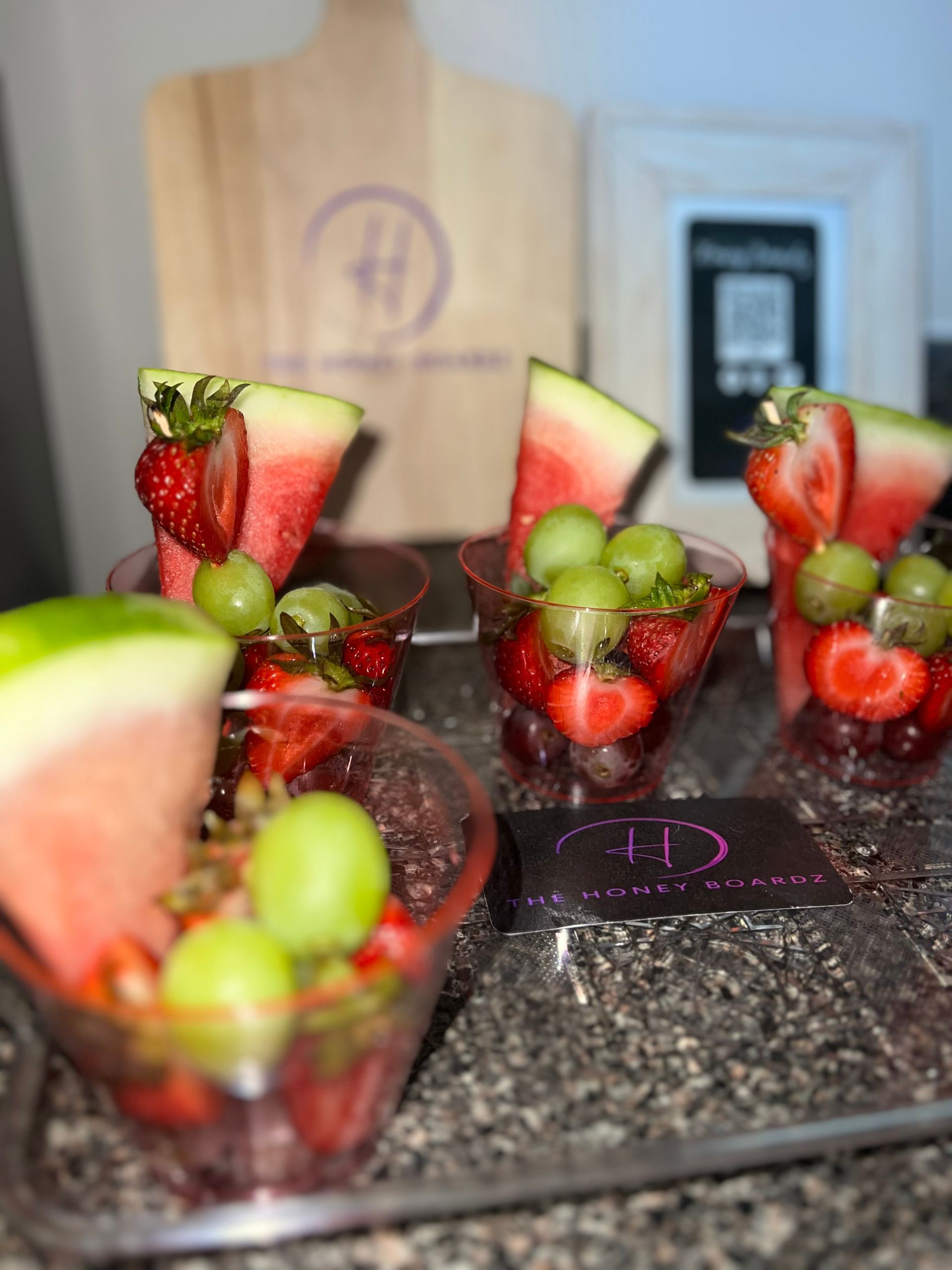 Fruit cups with strawberries and green grapes arranged on a countertop, with watermelon slices in the foreground.