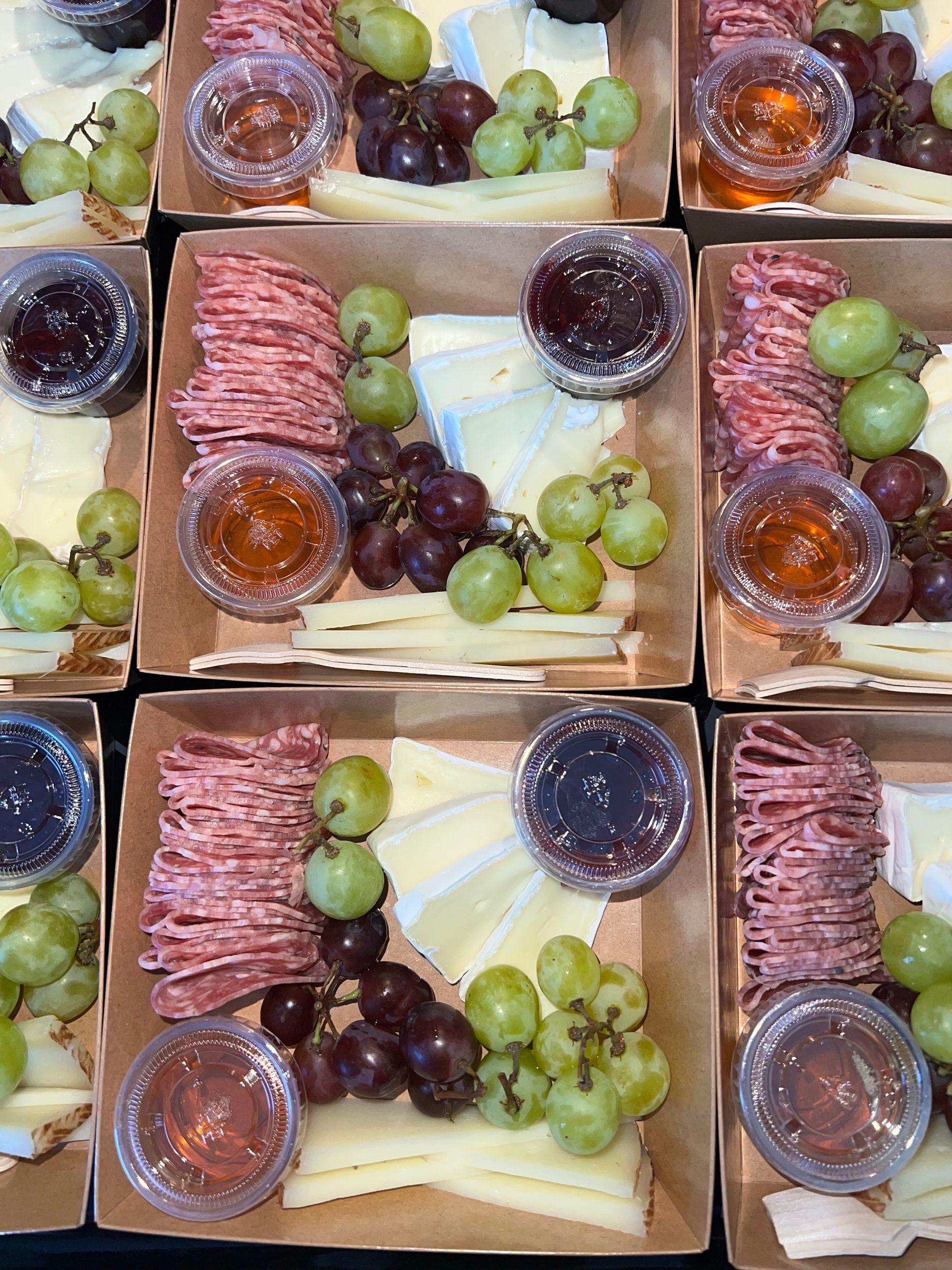 Three snack boxes with sliced meat, grapes, cheese, crackers, and small containers of dip or jelly.