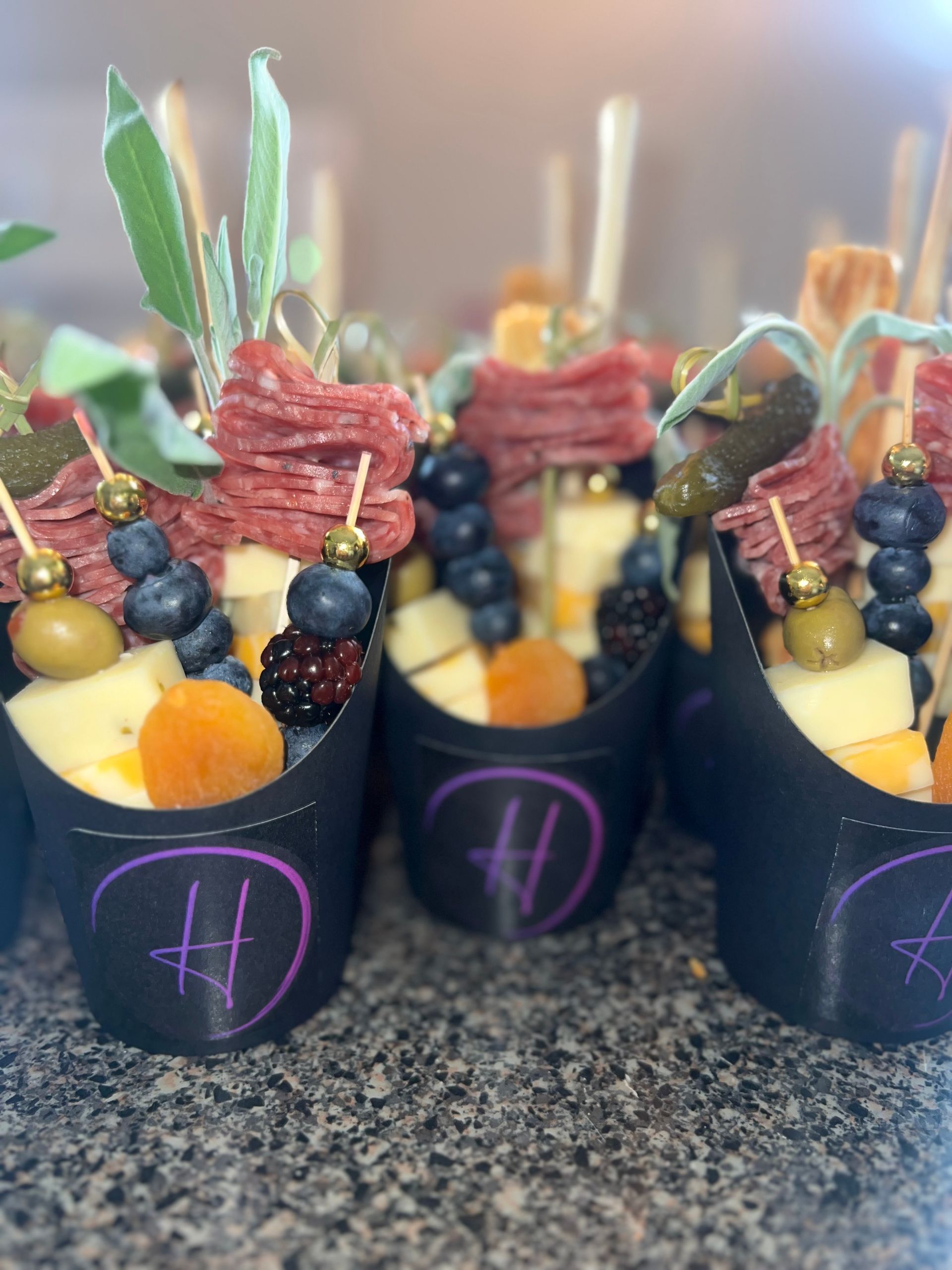Black dessert cups with fruit, cookies, and skewers on a countertop, arranged as party treats