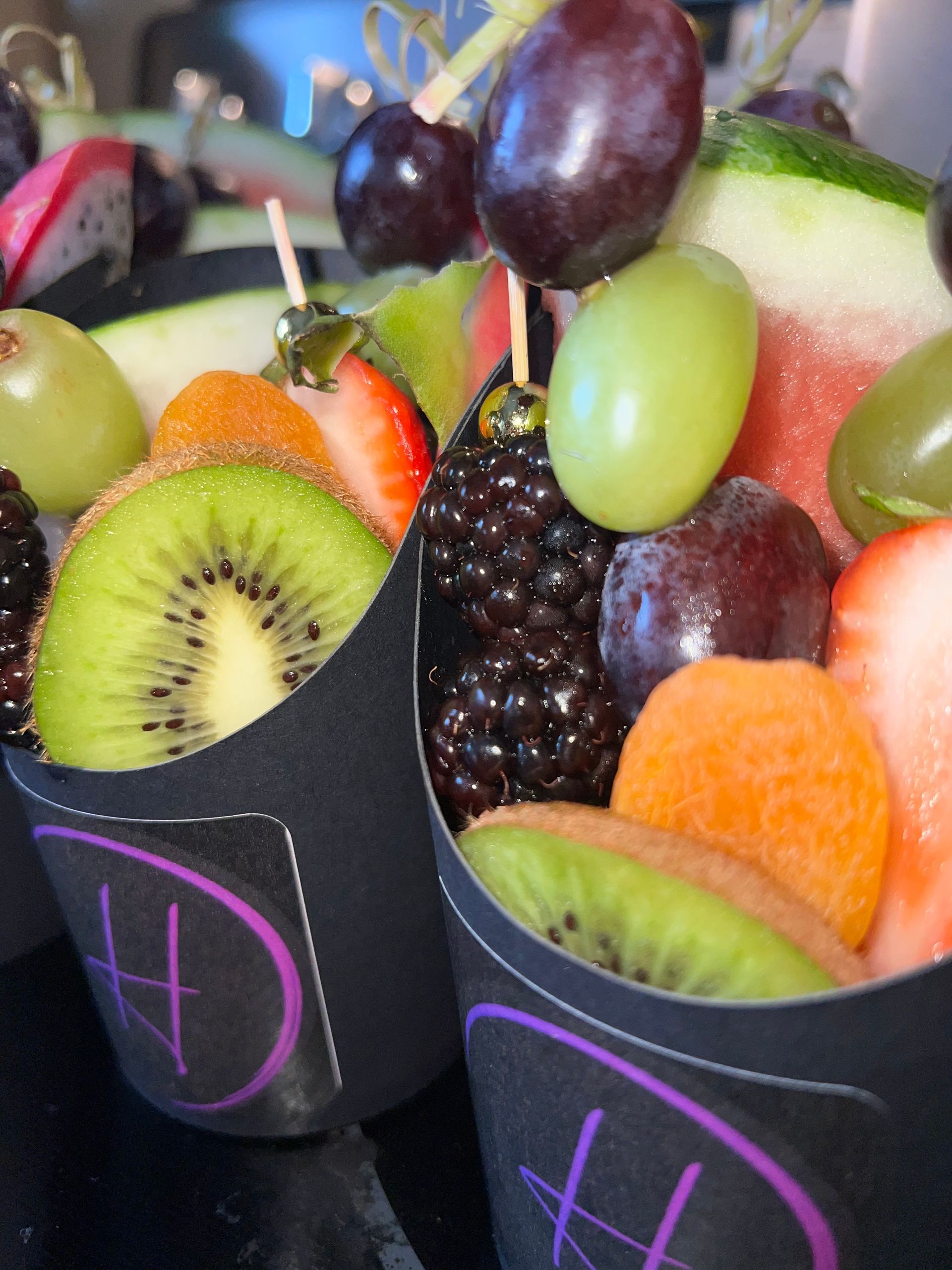 Colorful fruit cups with kiwi, grapes, berries, orange, and watermelon slices in black containers