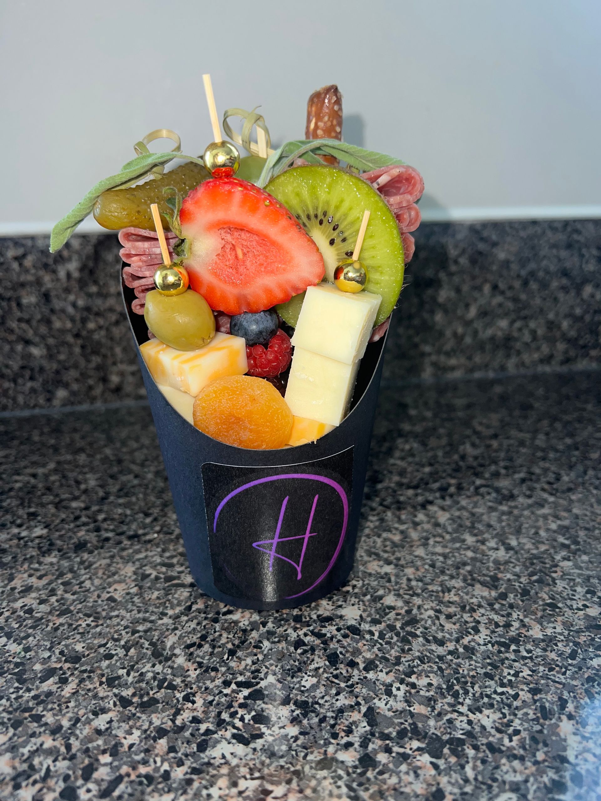 Fruit cup in a black cup, topped with strawberry, kiwi, orange, apple, and mint on a countertop