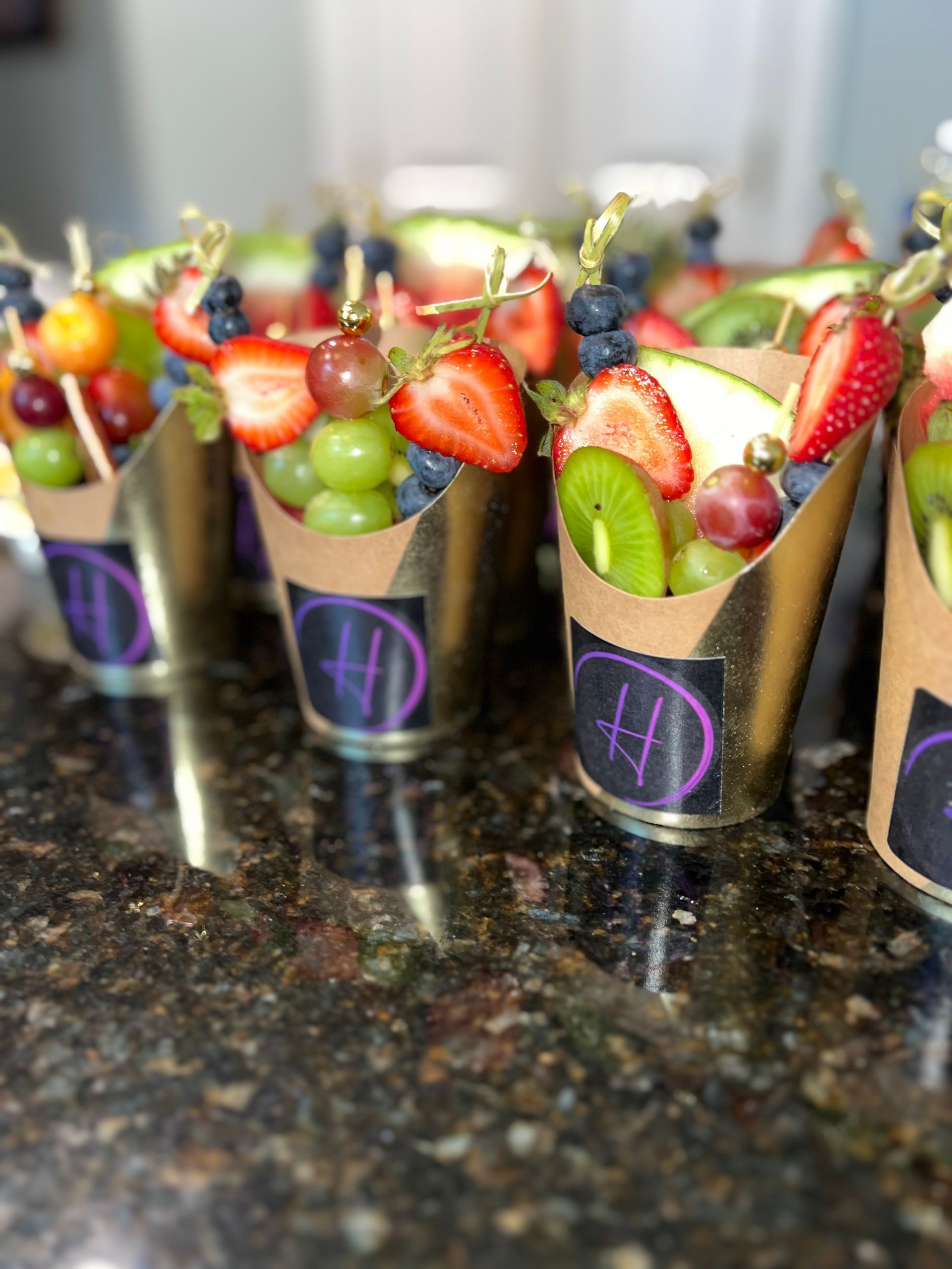 Fruit cups with strawberries, grapes, and kiwi in black paper holders on a countertop