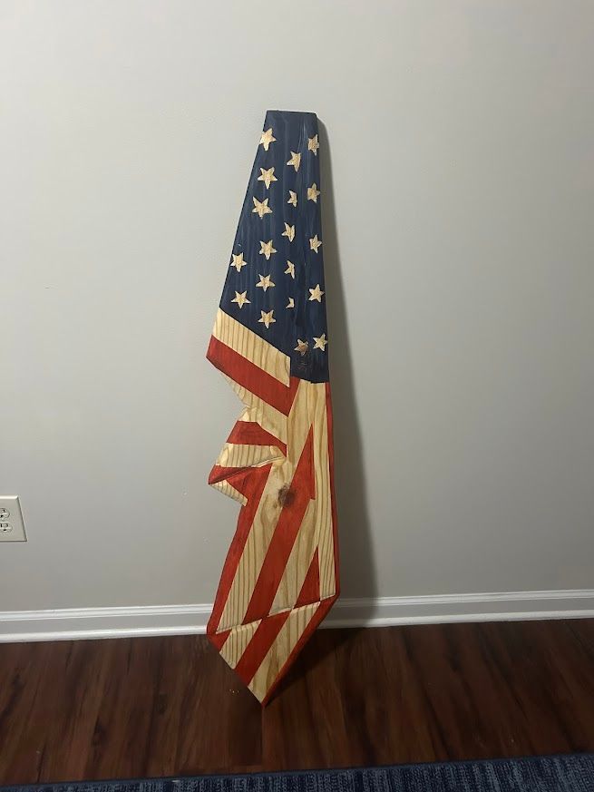 Wooden American flag wall hanging, partially folded. Red and white stripes, blue star field, leaning against a gray wall.