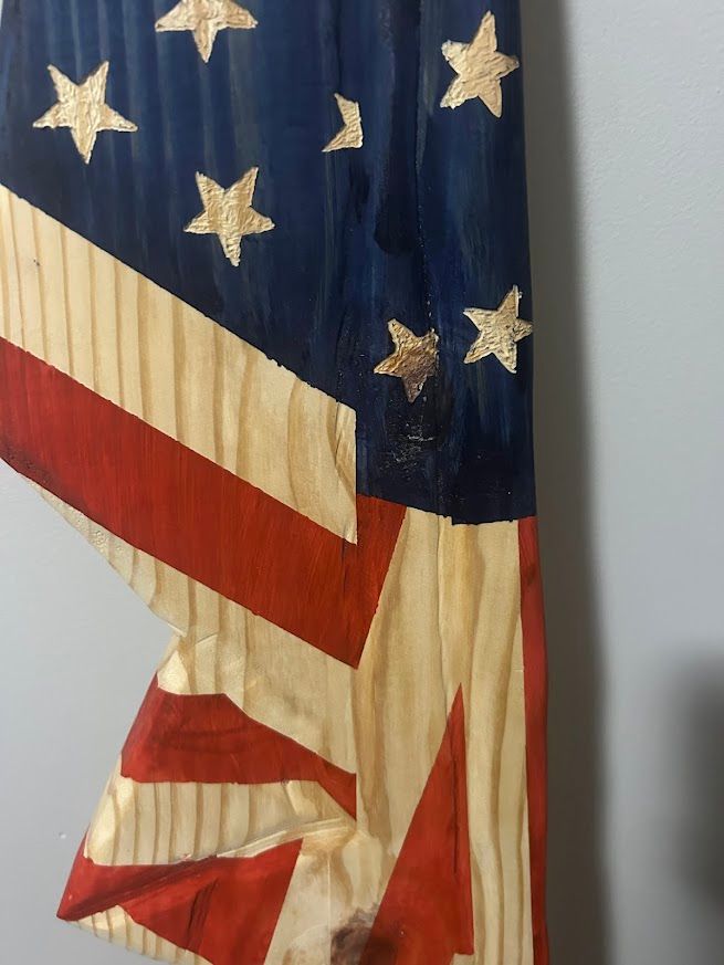 Wooden American flag wall hanging, partially folded. Red and white stripes, blue star field, leaning against a gray wall.