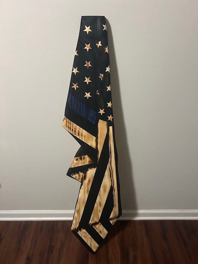 Wooden American flag with charred black and tan stripes, blue stripe, gold stars, leaning against a gray wall.