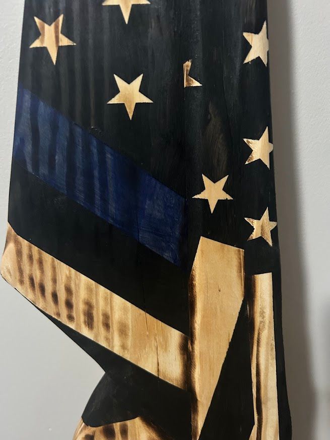 Wooden American flag with charred black and tan stripes, blue stripe, gold stars, leaning against a gray wall.