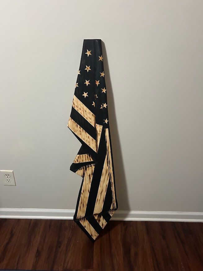 American flag, folded vertically, black with tan stripes and stars, mounted against a gray wall.