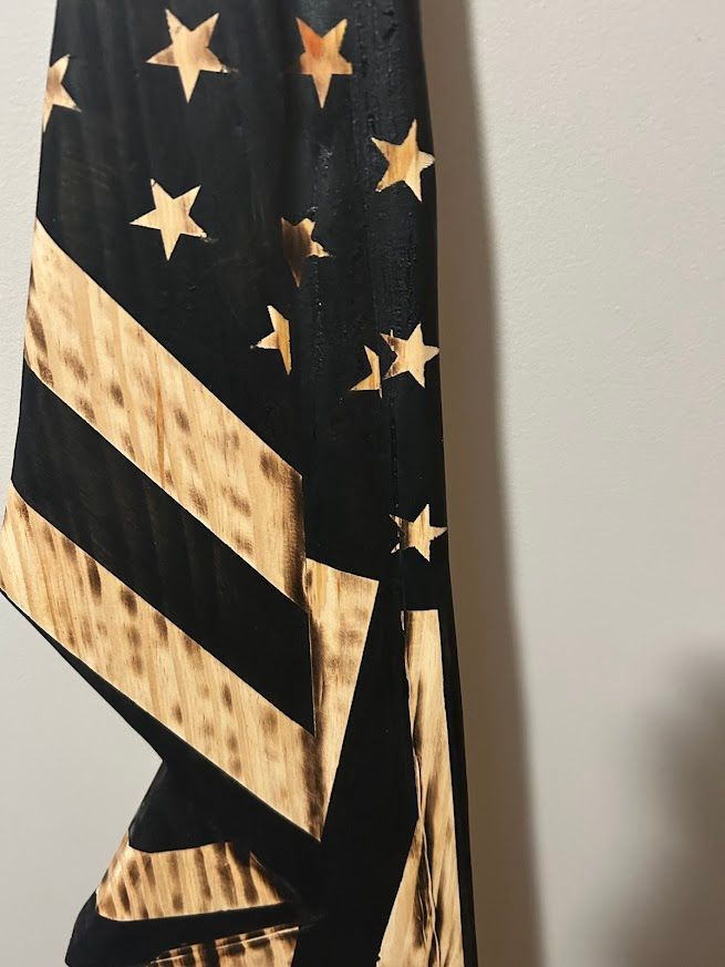 American flag, folded vertically, black with tan stripes and stars, mounted against a gray wall.