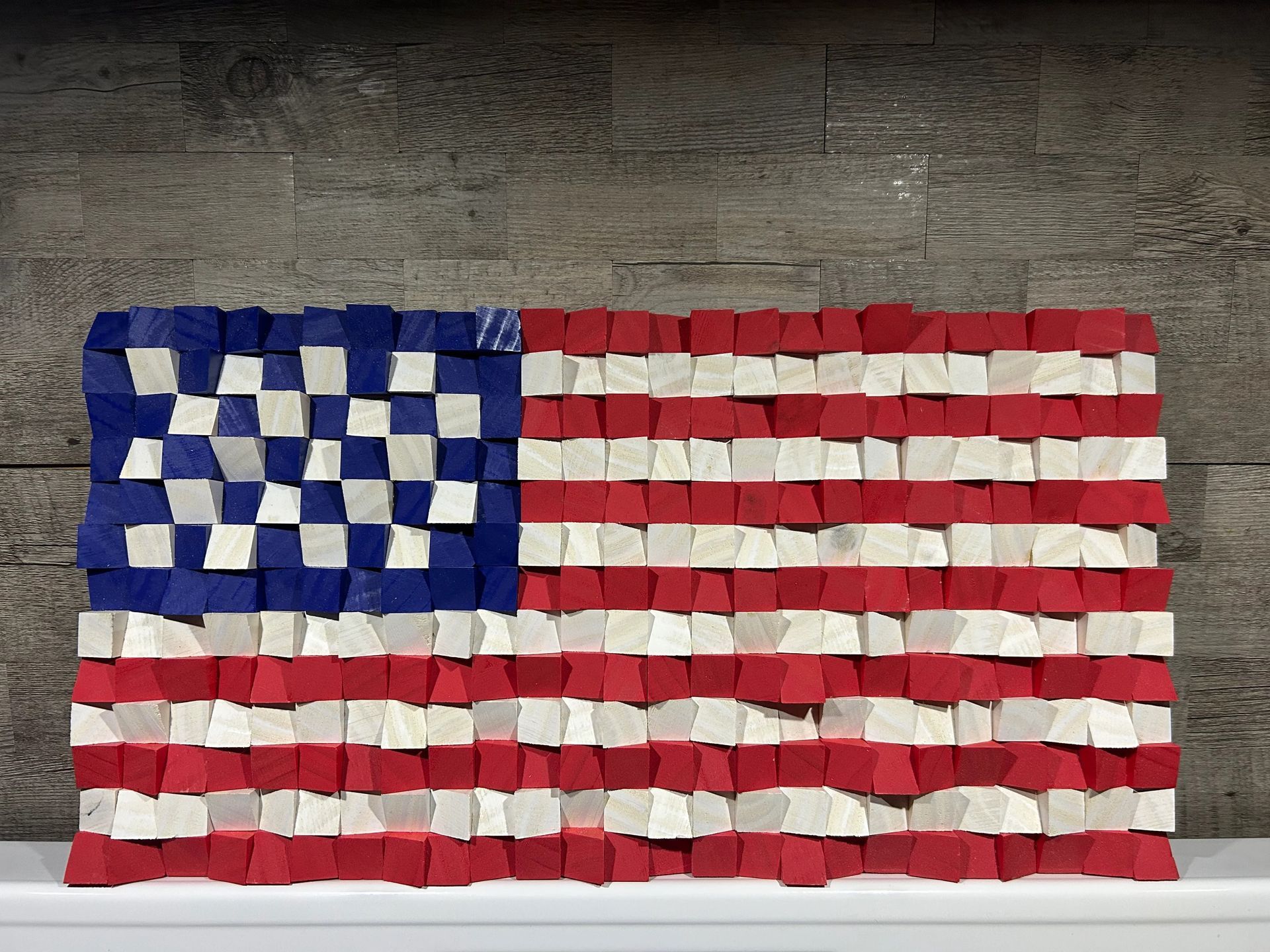 American flag art made from wooden blocks; red, white, and blue colors, displayed on a shelf against a wood wall.