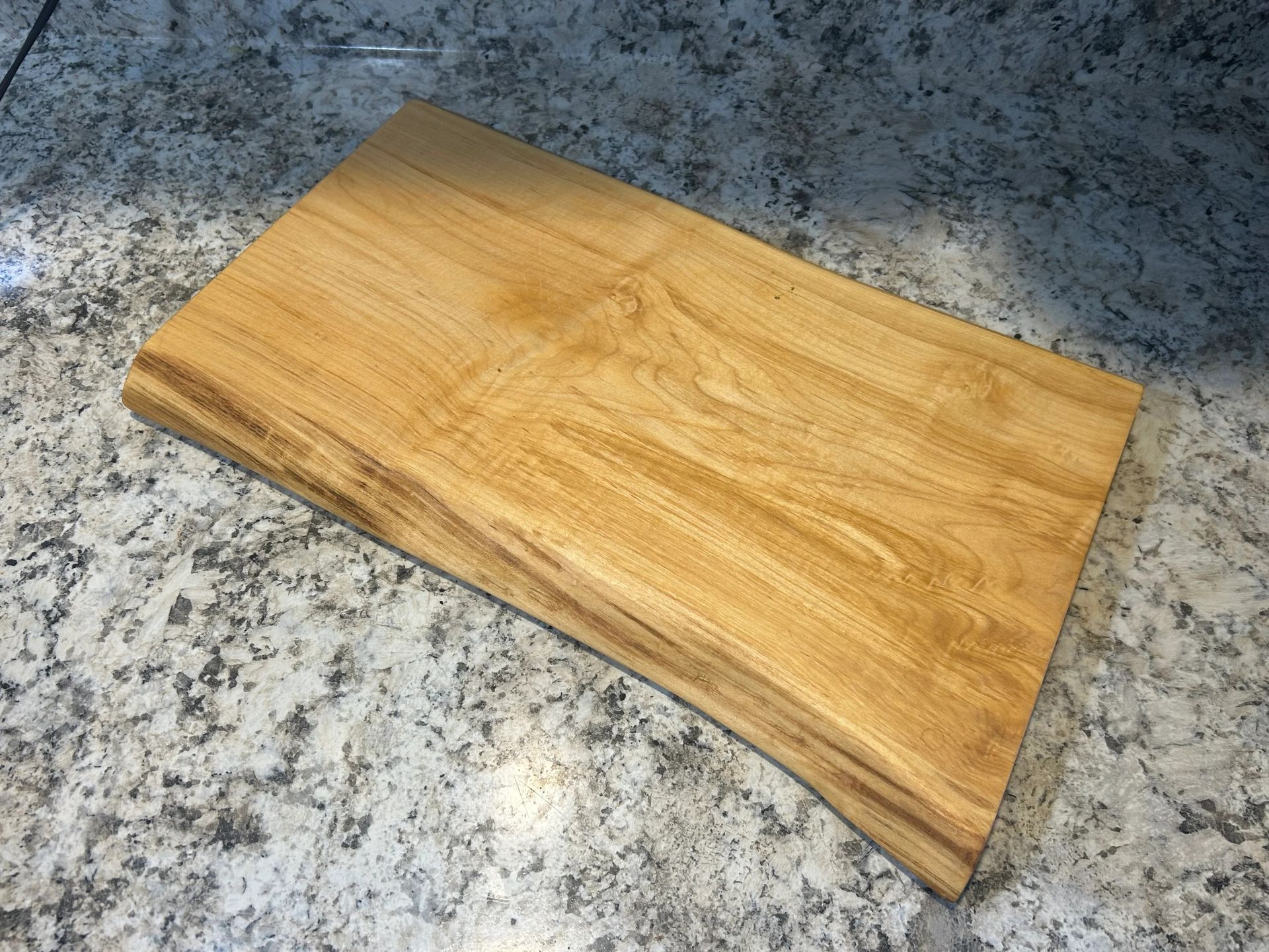Wooden cutting board with live edge, light brown color, resting on a speckled, gray countertop.