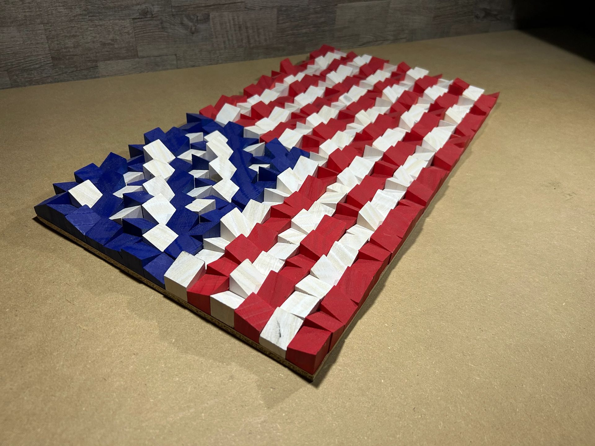 American flag art made from wooden blocks; red, white, and blue colors, displayed on a shelf against a wood wall.