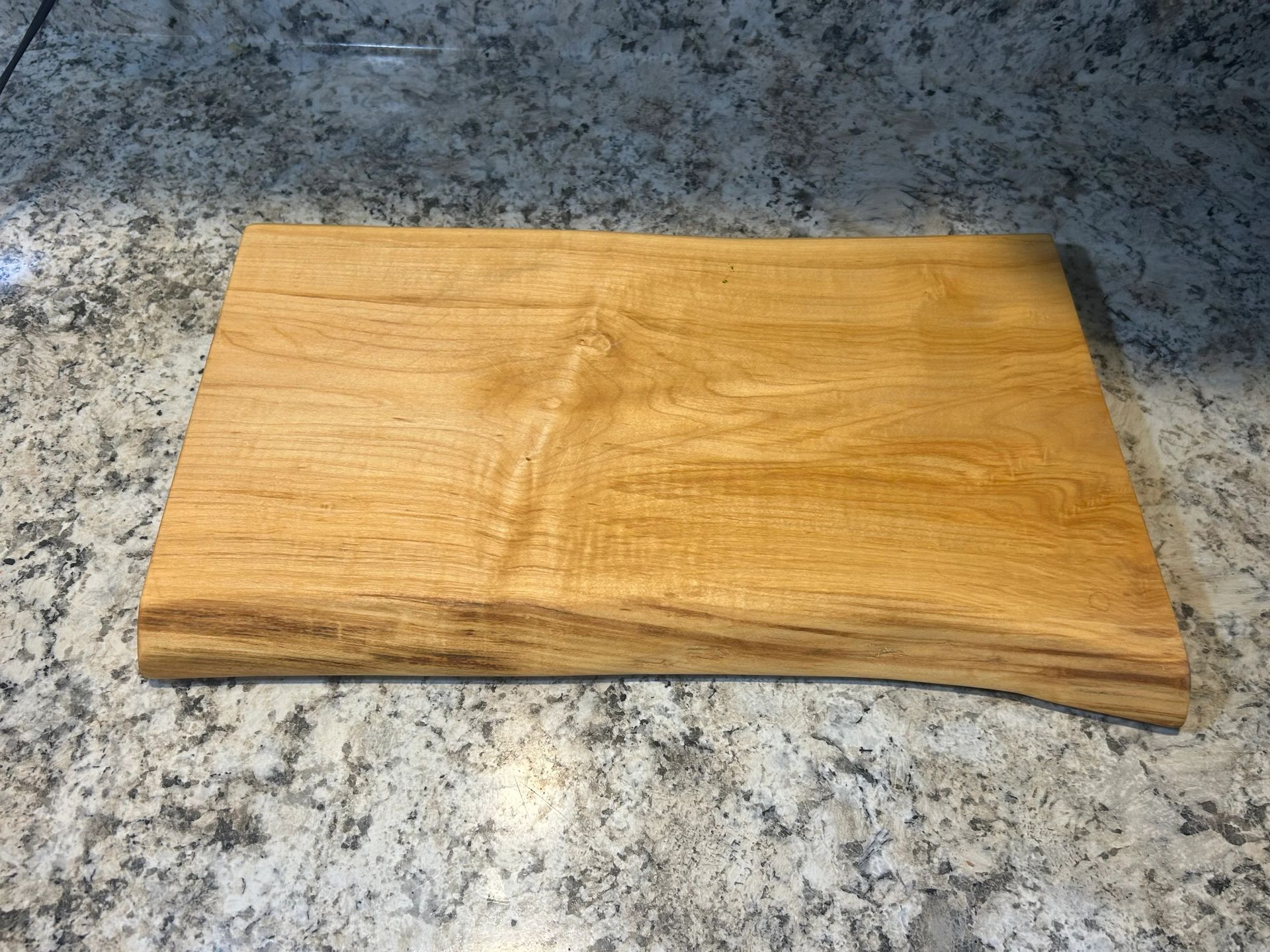 Wooden cutting board with live edge, light brown color, resting on a speckled, gray countertop.