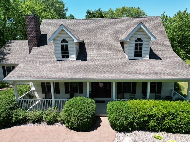 House with gray roof, white trim, front porch, two dormers, and surrounding green foliage.