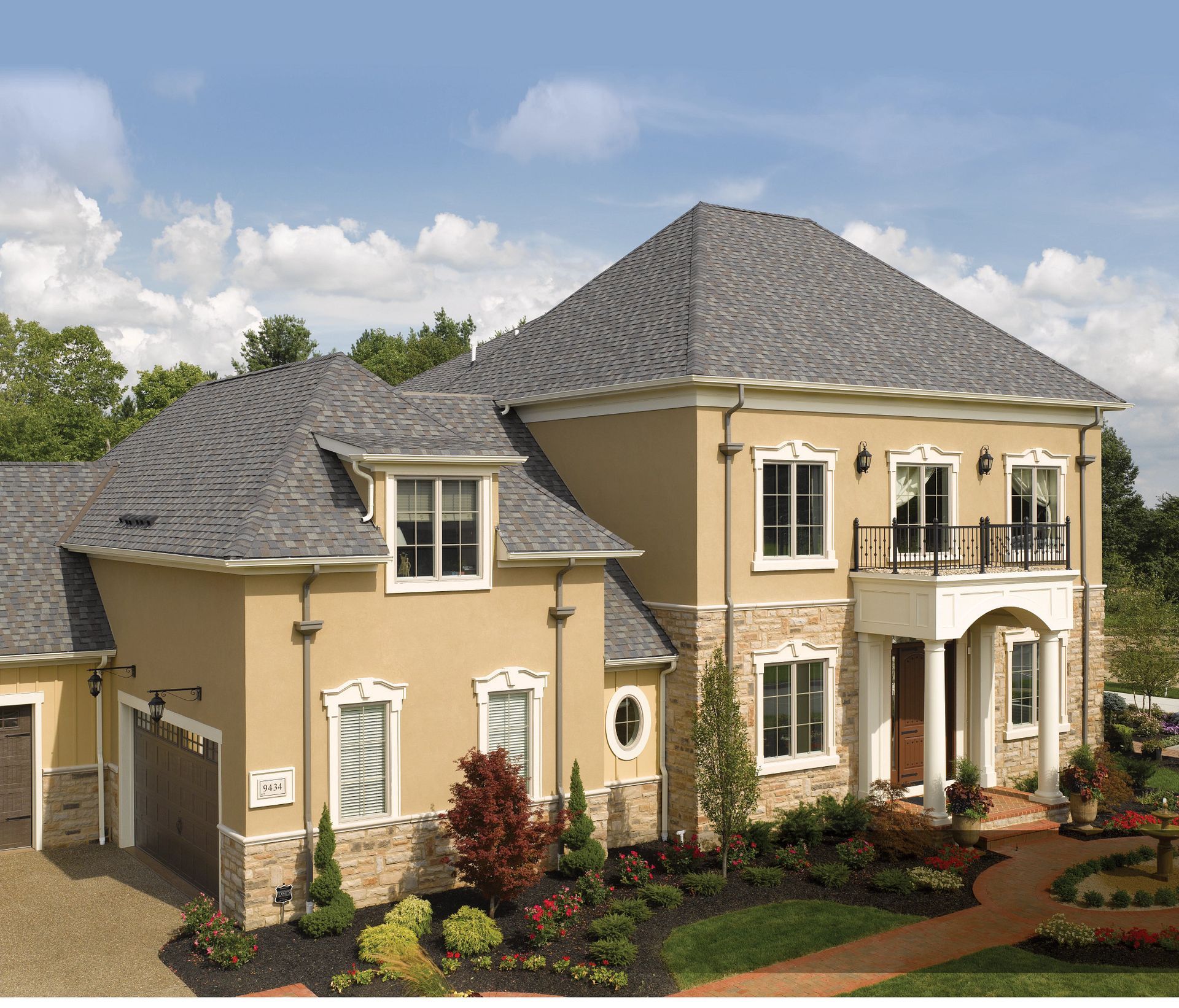 Two-story beige house with gray roof, white trim, stone accents, and landscaped yard under a blue sky.
