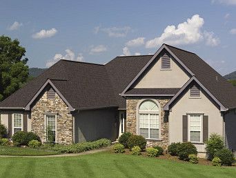 House with stone and tan facade, dark roof, green lawn, blue sky.