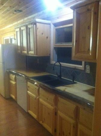 A rustic kitchen with wood-paneled walls and cabinets, a dark countertop, a double sink, and a white dishwasher.