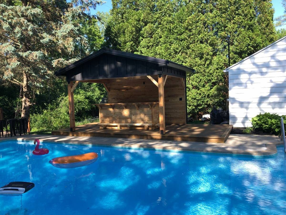 A wooden pool bar under a covered pavilion, adjacent to a bright blue swimming pool with floating inflatables.