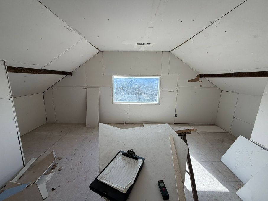 Unfinished attic room with white drywall, a small window, and construction materials on the floor.