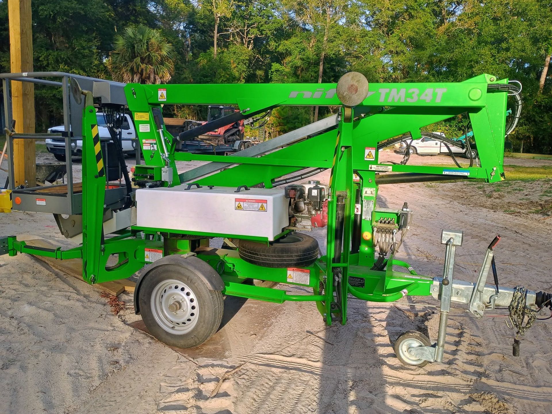 Green towable lift parked on a sandy path beside trees and equipment.