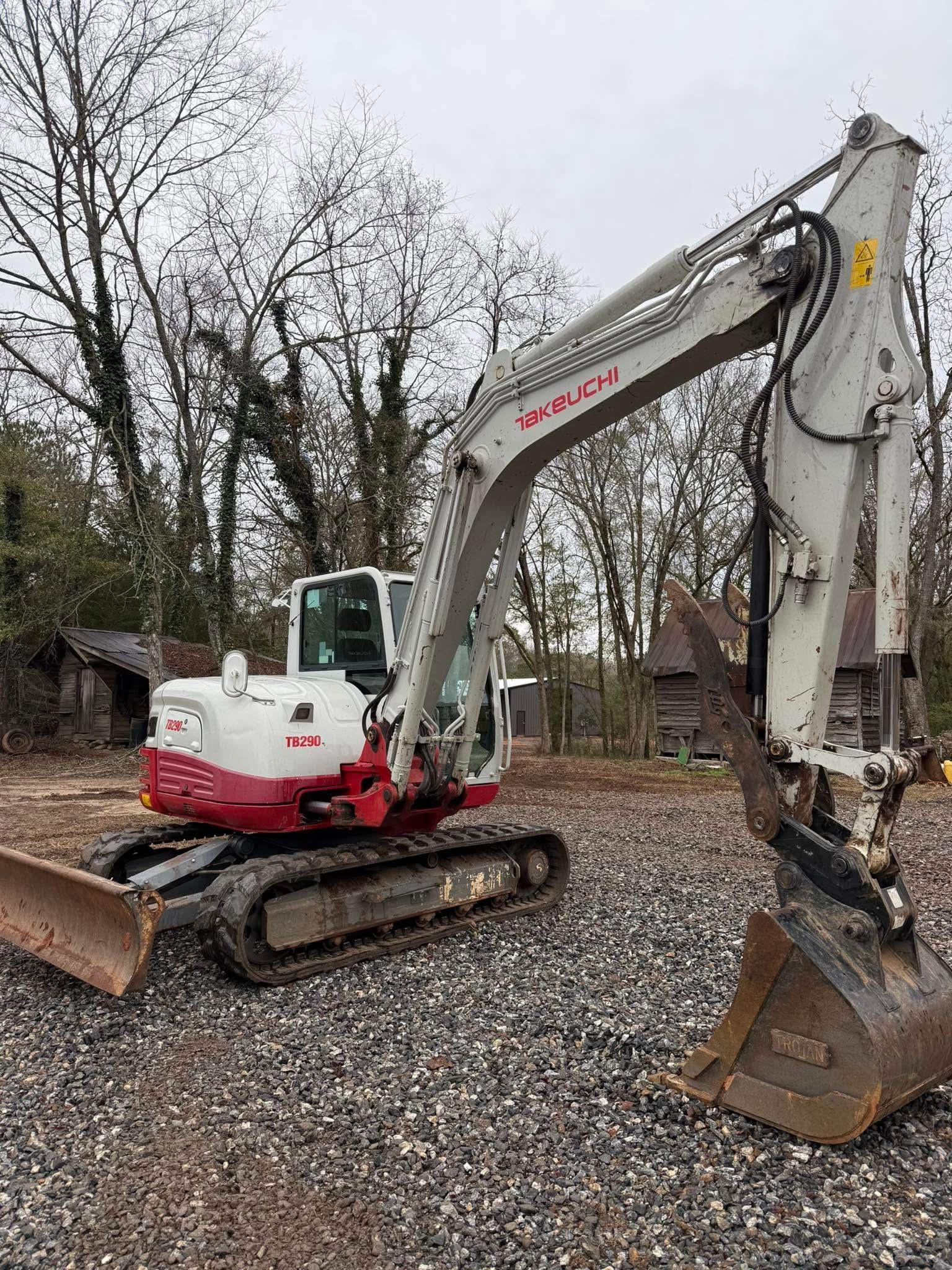 White and red excavator parked on gravel in a wooded area, bucket lowered to the ground