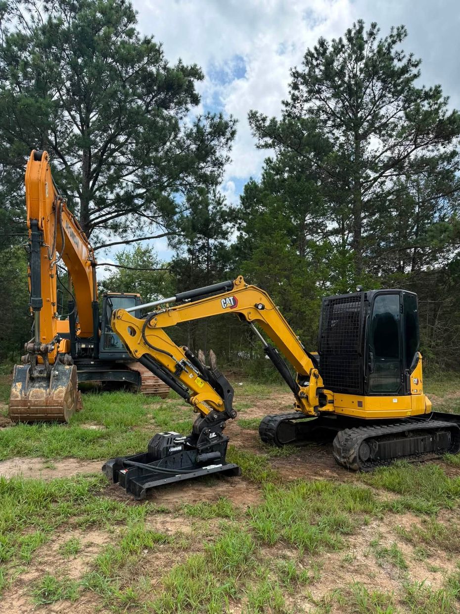 Yellow and black mini excavator on tracks beside a grassy dirt clearing with trees in the background