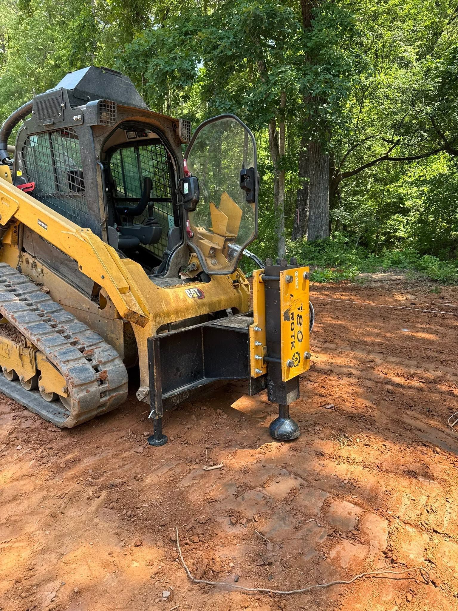 Yellow skid-steer loader with auger attachment on a dirt path in a wooded area