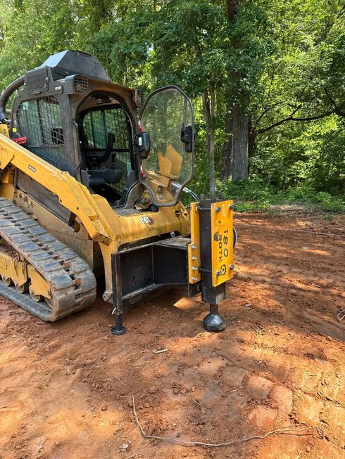 Yellow skid-steer loader with a front auger attachment on a muddy wooded site