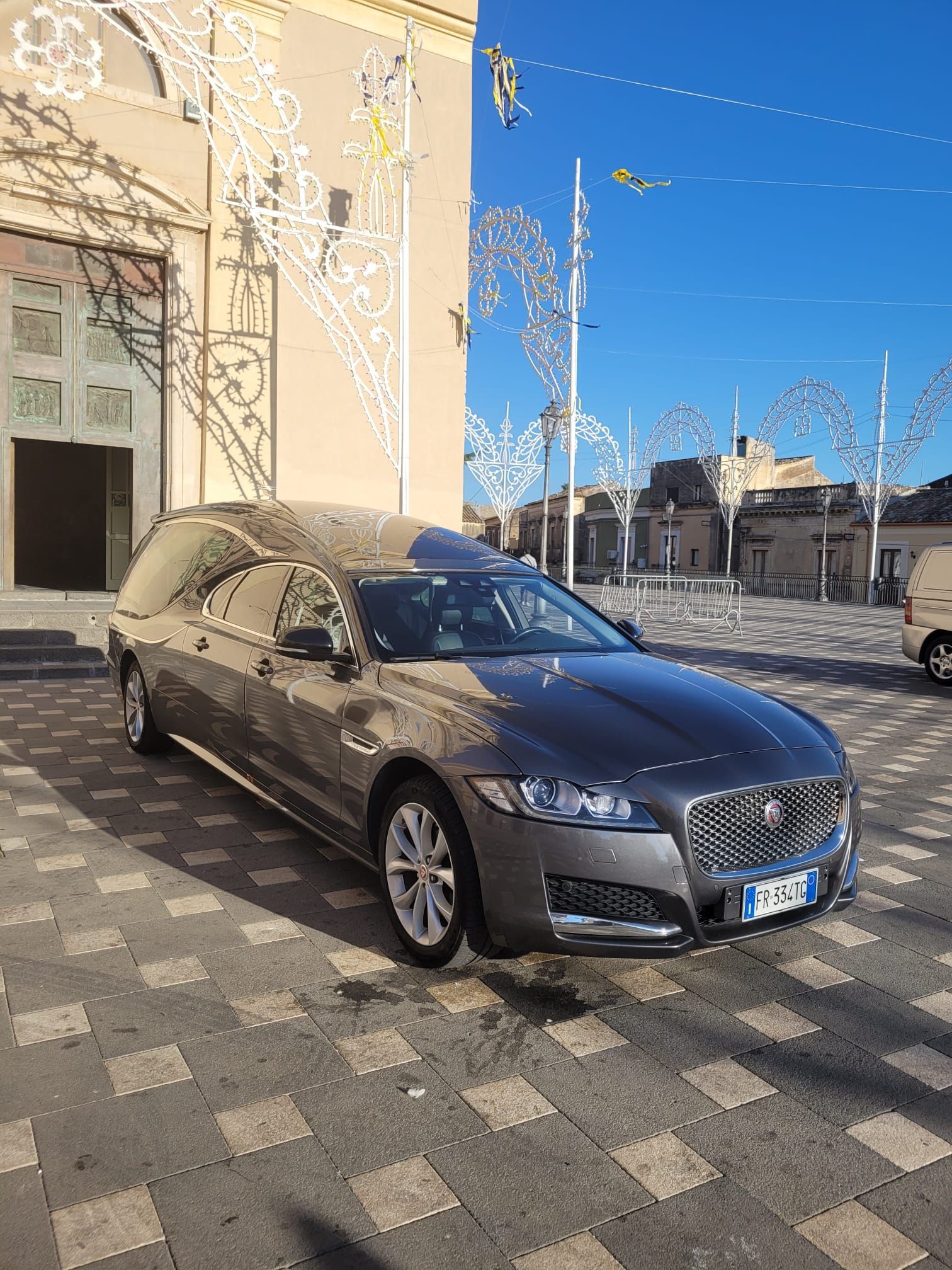 Carro funebre Jaguar grigio parcheggiato fuori da un edificio con luci natalizie; terreno acciottolato.