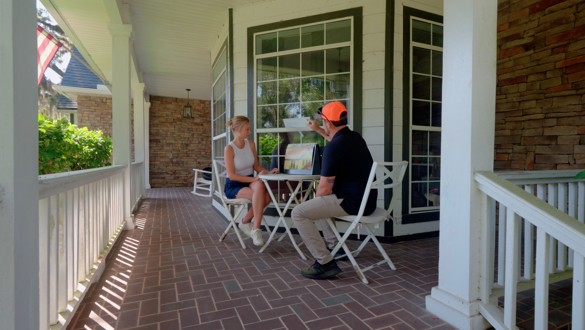 Two people sitting at a table on a covered brick porch, talking beside white railings and a window