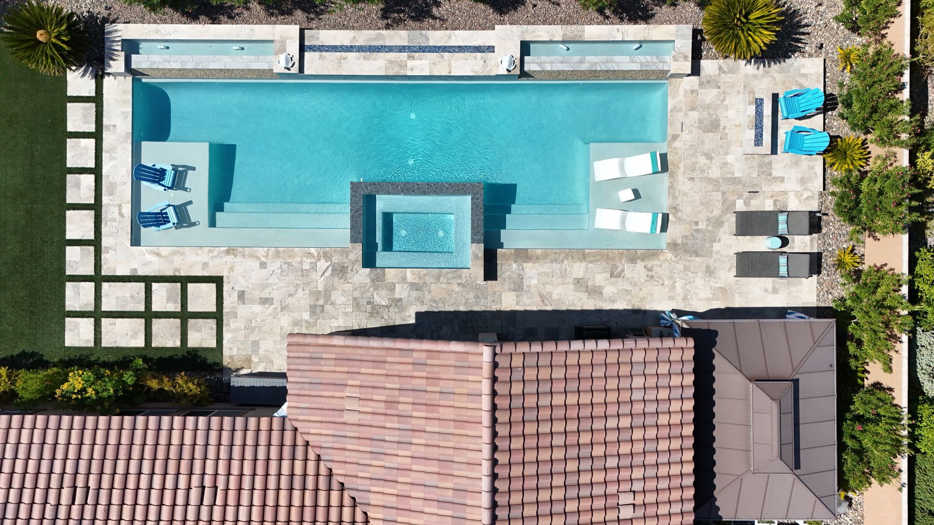 An aerial view of a rectangular backyard pool with a centered spa, sun shelves, lounge chairs, and a paved patio area.