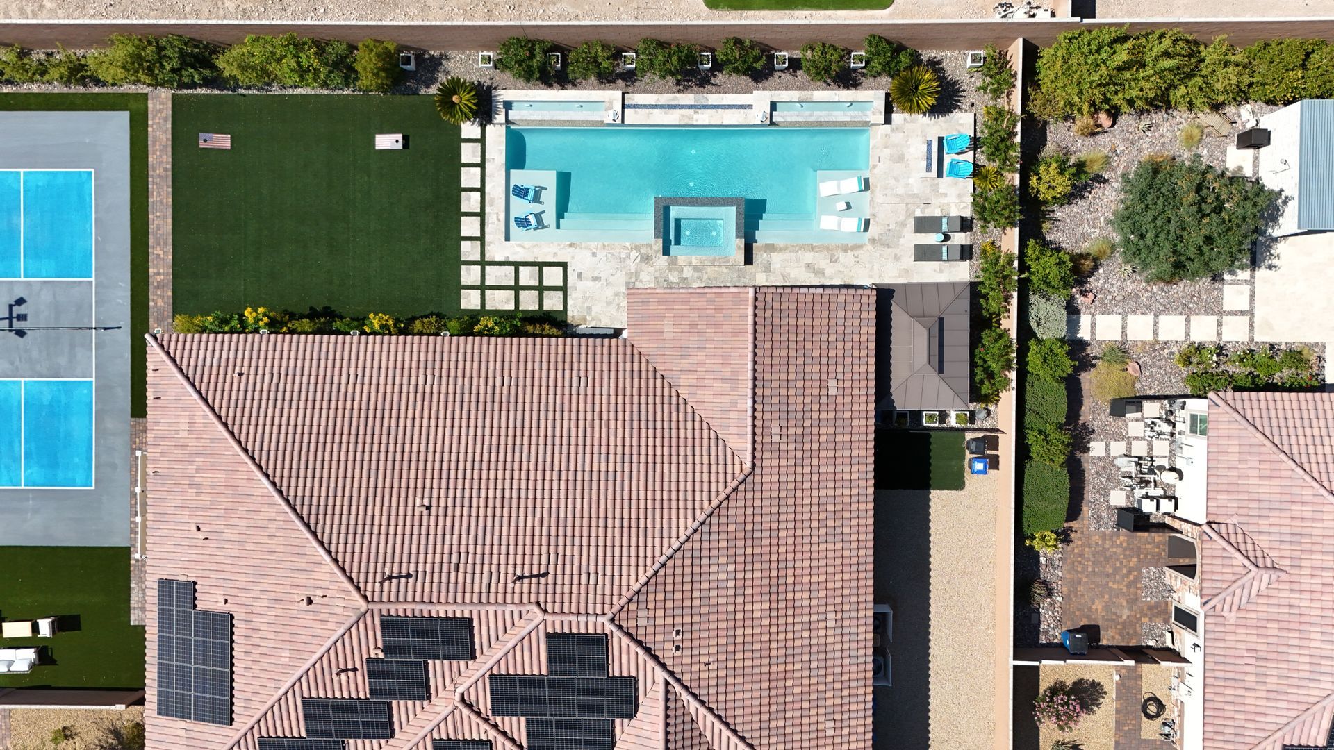 Aerial view of a home with a red-tiled roof, solar panels, a rectangular swimming pool, and a backyard pickleball court.